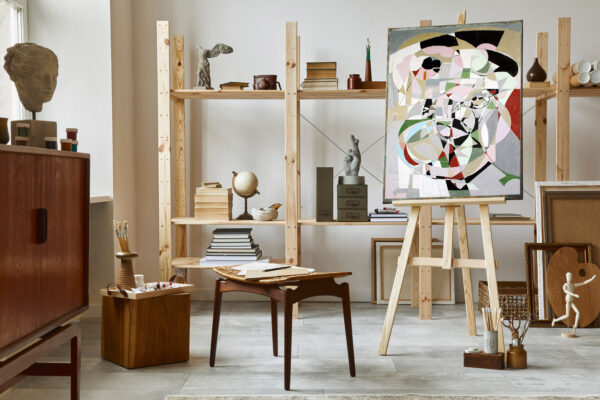 Unique artist workspace interior with stylish teak commode, wooden easel, bookcase, artworks, painting accessories, decoration and elegant personal stuff. Modern work room for artist. Template.
